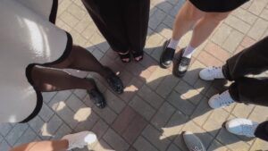 A group of people stands in a circle on a sunlit paved ground. Only their feet and legs are visible, showcasing diverse and stylish footwear, including black heels, loafers, and white sneakers. The ground consists of gray and brown bricks forming a pattern.