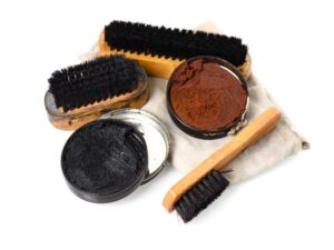 Shoe polishing supplies are displayed, featuring two open tins of polish—one black and one brown. Highlighted are three shoe brushes of varying sizes, each with black bristles, set atop a cloth. Choosing the right brush type complements this collection, all elegantly arranged against a white background.