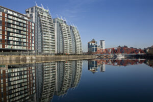 A row of modern apartment buildings with distinctive curved glass facades reflect on the calm waters of a canal in Manchester. A red metal bridge crosses the canal, offering visitors unique travel experiences, and more buildings are visible in the background under a clear blue sky.